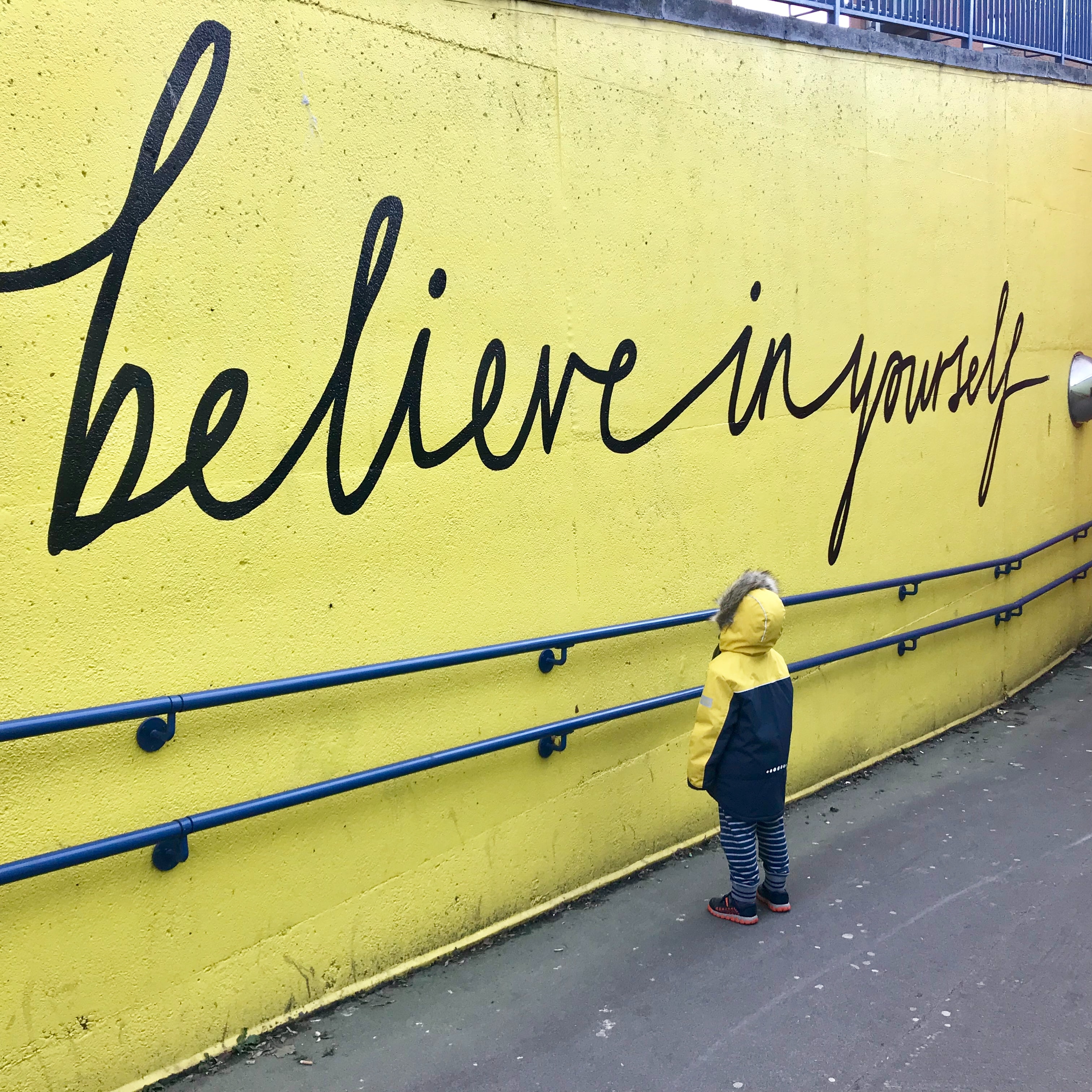 kid in jacket looking at yellow wall with believe in yourself written